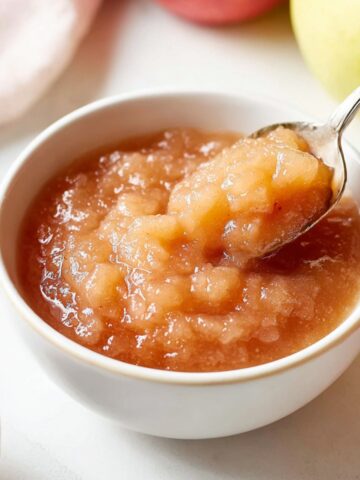 Bowl of homemade crockpot applesauce with a spoon lifting a chunky scoop, surrounded by fresh apples on a bright kitchen counter.