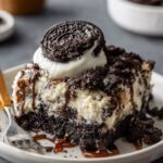 A slice of homemade Oreo Dump Cake topped with whipped cream, an Oreo cookie, and a drizzle of caramel, served on a rustic plate with a fork beside it.