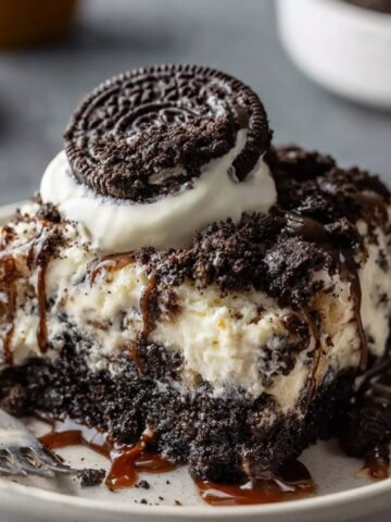 A slice of homemade Oreo Dump Cake topped with whipped cream, an Oreo cookie, and a drizzle of caramel, served on a rustic plate with a fork beside it.