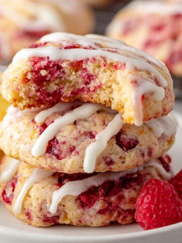 Homemade Raspberry Lemon Cookies with white icing, one bitten into, placed on a white plate with fresh raspberries and a lemon wedge.