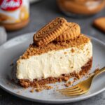Slice of Lotus Biscoff Cheesecake topped with whole biscuits and cookie crumbs, with a gold fork beside it.