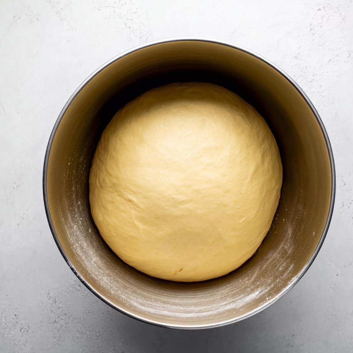 A bowl of risen yeast dough sits in a metal mixing bowl on a light, textured countertop.