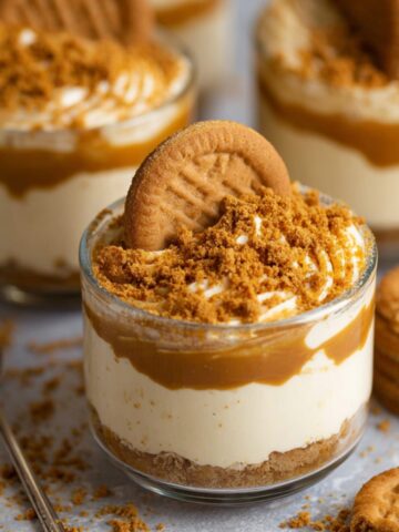 No-Bake Biscoff Cheesecake Cups topped with whipped cream, caramel sauce, crushed Biscoff cookies, and a whole Biscoff biscuit.