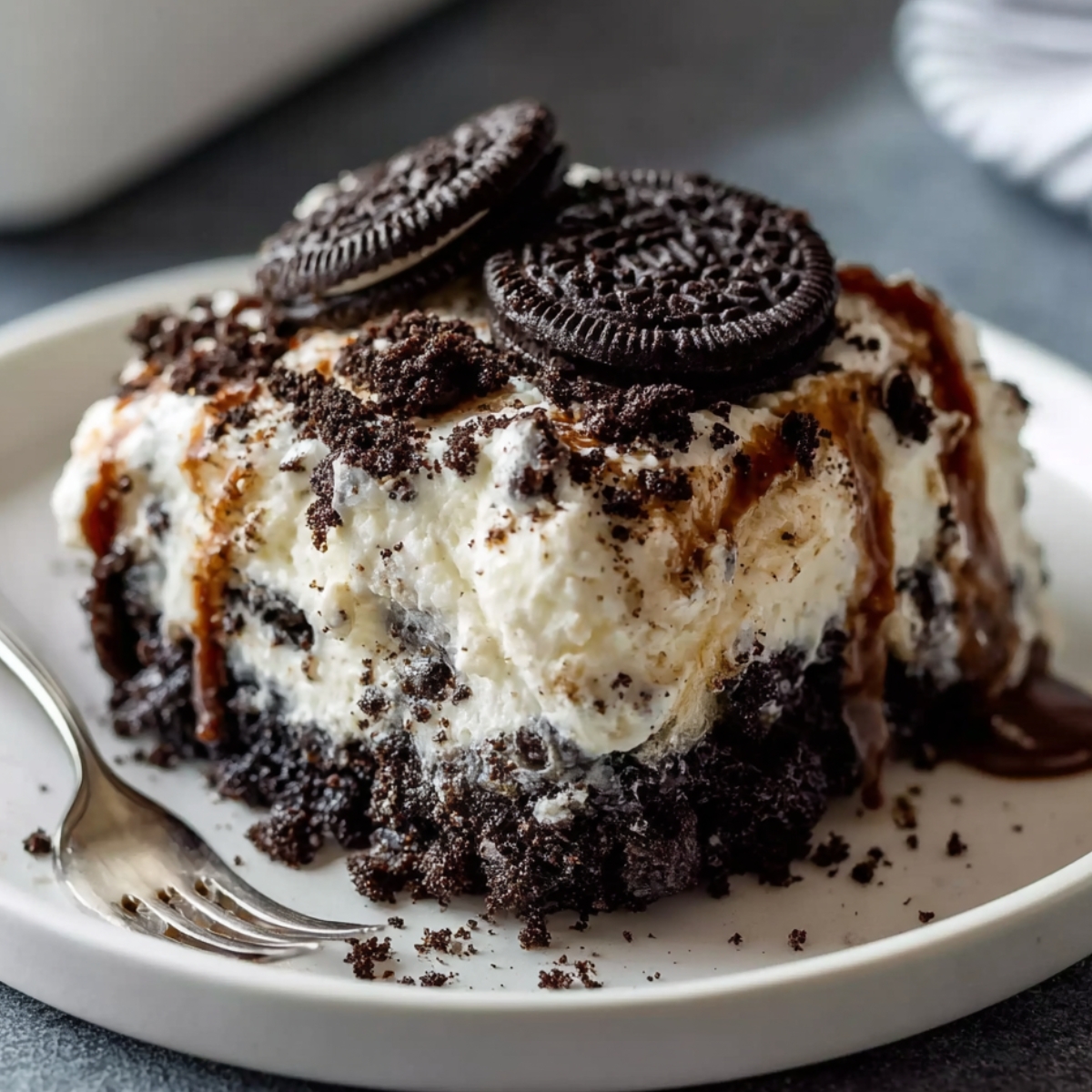Easy Oreo Dump Cake Recipe 11 A slice of homemade Oreo Dump Cake with whipped cream, a whole Oreo cookie on top, and caramel drizzle, served on a rustic plate with a fork beside it.