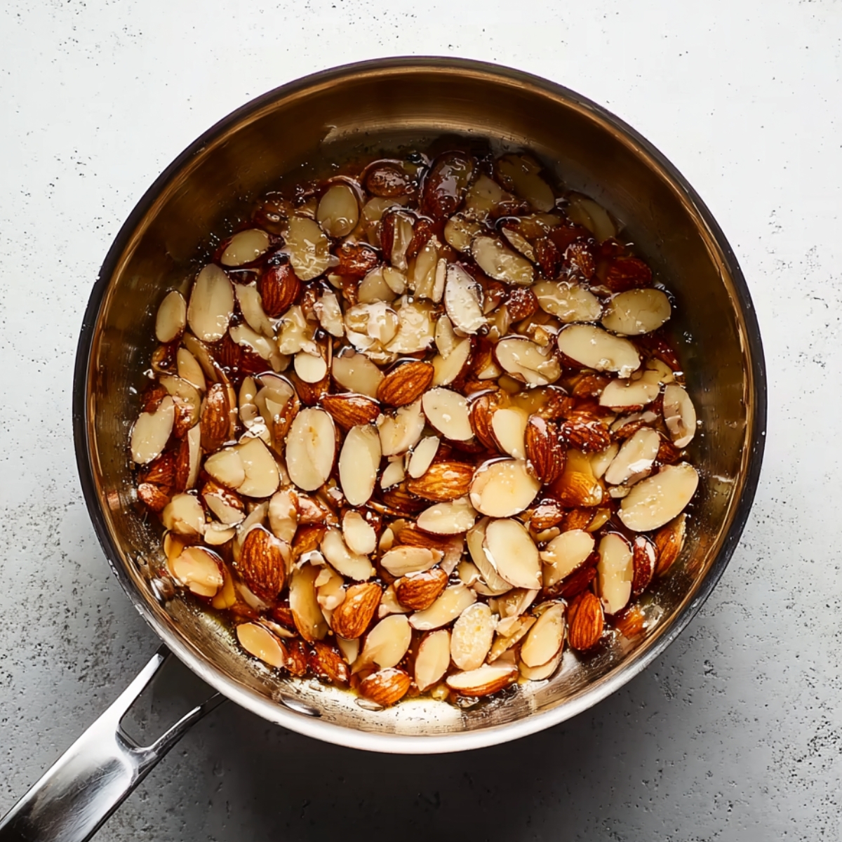 A saucepan filled with sliced almonds coated in a glossy caramel mixture sits on a light textured countertop.