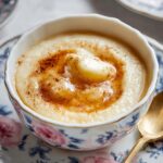 A bowl of creamy Rømmegrøt topped with a square of butter, a drizzle of syrup, and a sprinkle of cinnamon, served on a floral plate with a golden spoon.