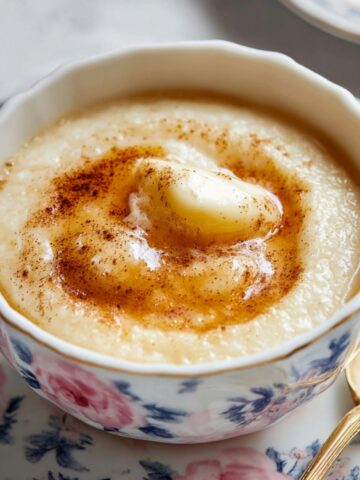 A bowl of creamy Rømmegrøt topped with a square of butter, a drizzle of syrup, and a sprinkle of cinnamon, served on a floral plate with a golden spoon.