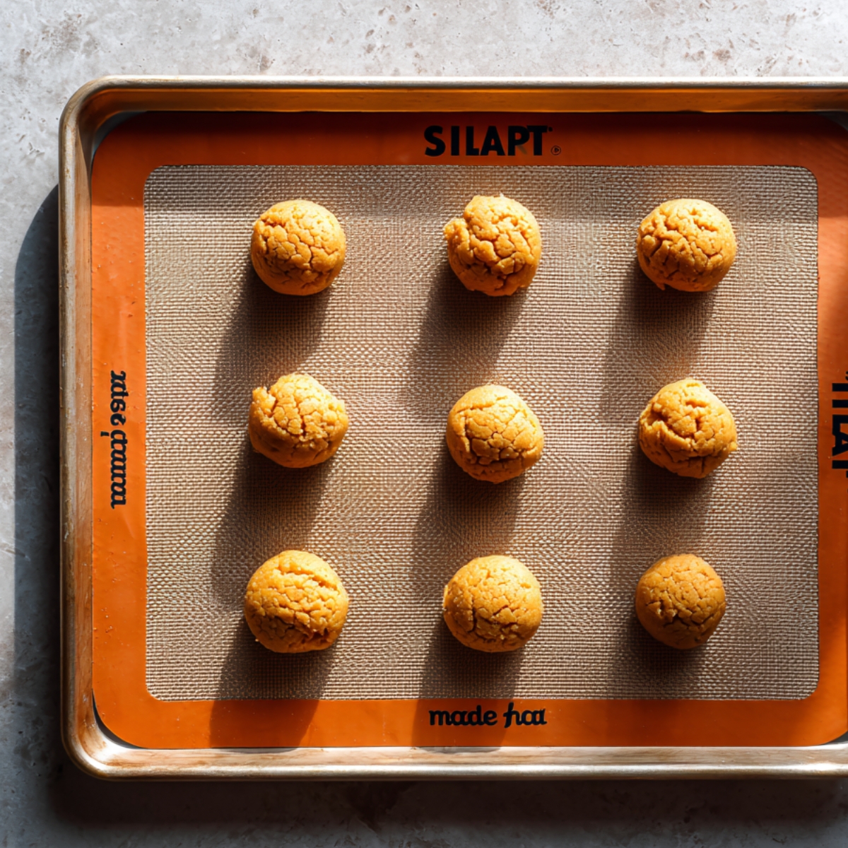 Pumpkin cookie dough balls arranged on a baking sheet lined with a Silpat mat, ready to be baked.
