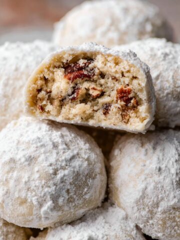 A stack of Snowball Cookies dusted with powdered sugar, with one halved to reveal a pecan-filled, crumbly interior.