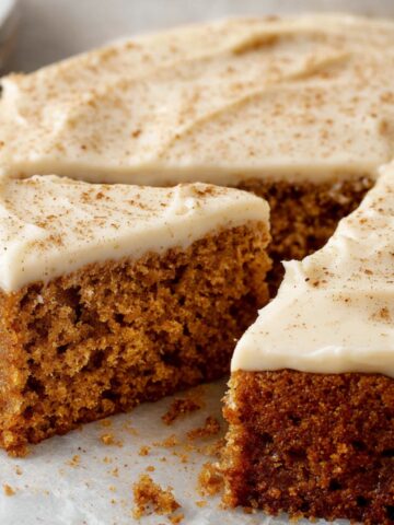 A homemade spice cake with cream cheese frosting, lightly dusted with cinnamon. A slice has been cut, revealing the moist, tender crumb inside. Crumbs are scattered on the parchment paper, giving it a freshly baked look.