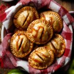 A basket filled with golden brown Apple Hand Pies resting on a red and white checkered cloth, each pie crimped around the edges and drizzled with caramel sauce, with fresh green apples and a rustic wooden table in the background.