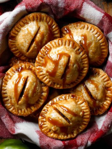 A basket filled with golden brown Apple Hand Pies resting on a red and white checkered cloth, each pie crimped around the edges and drizzled with caramel sauce, with fresh green apples and a rustic wooden table in the background.