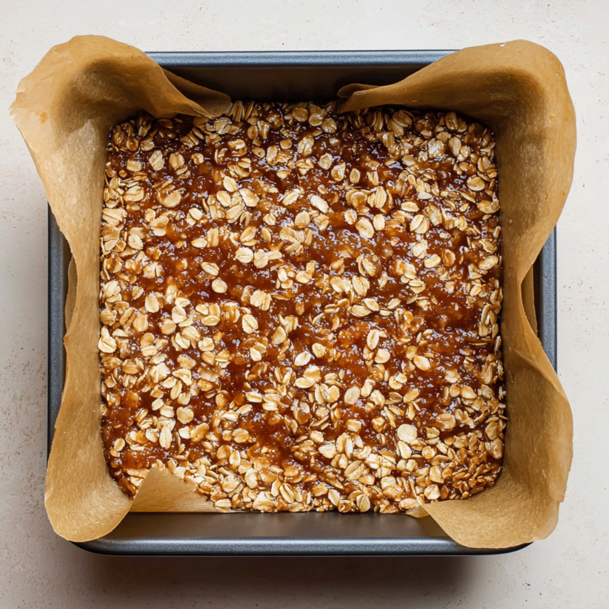 Homemade Oatmeal Fig Bars Recipe | Easy & Healthy 12 Square baking pan lined with parchment paper, filled with a mixture of oats and fig filling. The oats are spread evenly over the sticky fig layer, creating a textured topping ready for baking.