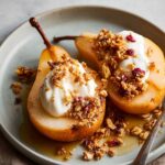 Two baked pears topped with yogurt, granola, and dried cranberries, served on a light plate with a drizzle of syrup.