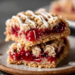 Close-up of two stacked Cherry Pie Bars, with a sweet glaze drizzled on top. The bars have a crumbly, golden oat crust, filled with vibrant red cherry filling.