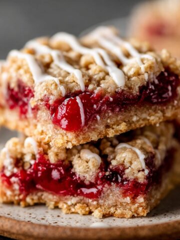 Close-up of two stacked Cherry Pie Bars, with a sweet glaze drizzled on top. The bars have a crumbly, golden oat crust, filled with vibrant red cherry filling.