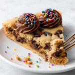 Slice of Chocolate Chip Cookie Cake topped with chocolate frosting and rainbow sprinkles on a white plate