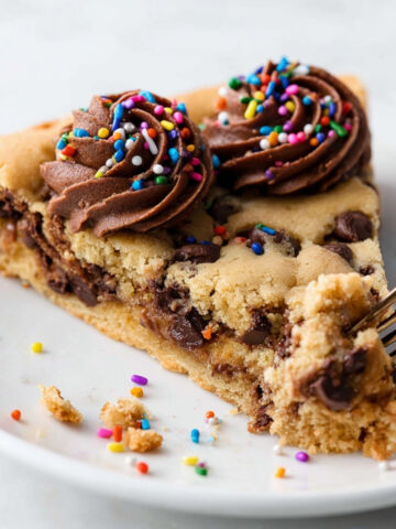 Chocolate Chip Cookie Cake Recipe | Easy & Chewy 17 Slice of Chocolate Chip Cookie Cake topped with chocolate frosting and rainbow sprinkles on a white plate