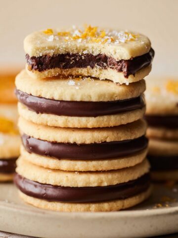 A stack of buttery Chocolate Orange Sandwich Cookies Recipe filled with smooth chocolate ganache, arranged on a ceramic plate and sprinkled with sugar and orange zest in soft natural light.