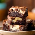 Close-up view of thick, moist Coconut Cheesecake Brownies stacked on a plate, with visible chunks of chocolate and creamy cheesecake layers inside