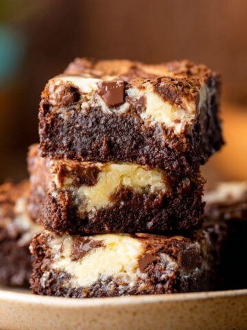 Close-up view of thick, moist Coconut Cheesecake Brownies stacked on a plate, with visible chunks of chocolate and creamy cheesecake layers inside