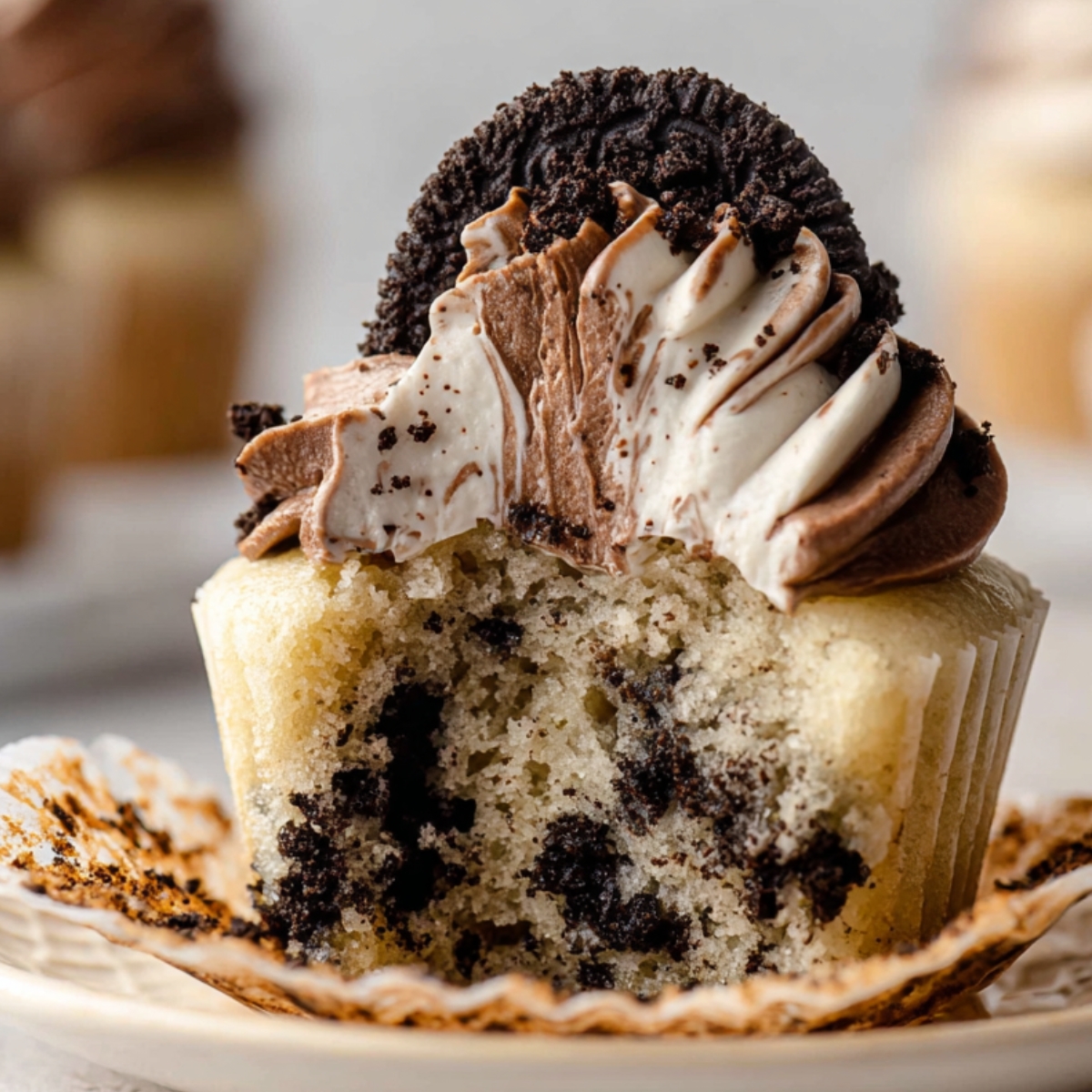 A Cookies and Cream Cupcakes with chocolate and vanilla frosting swirls topped with an Oreo cookie, showing a bite taken out of the cupcake.