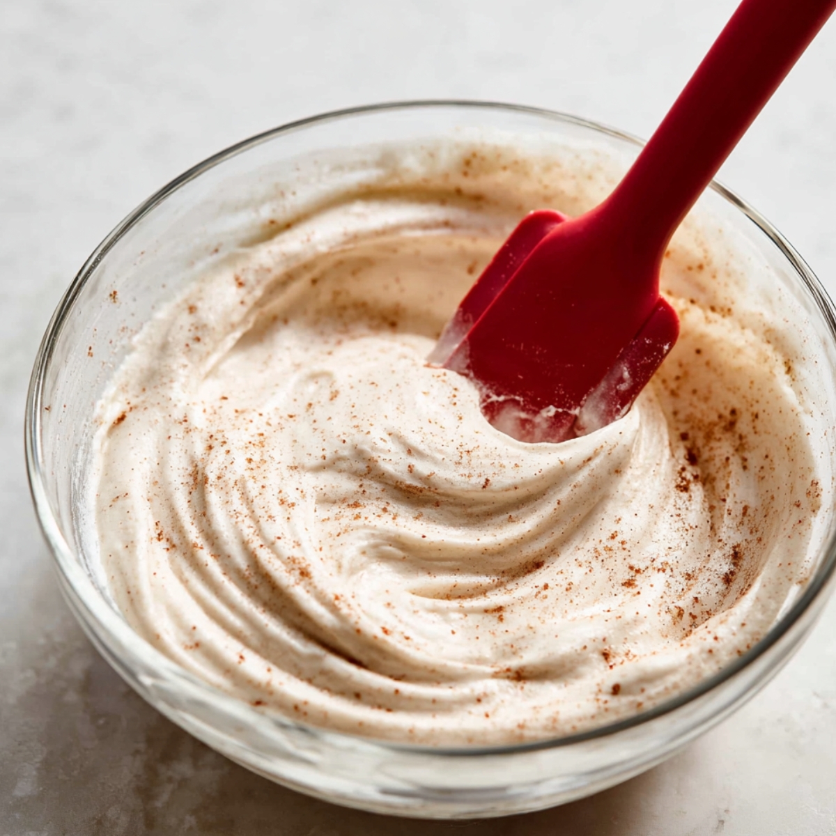 Hummingbird Bundt Cake Recipe | Moist & Easy 12 Cinnamon-spiced cream cheese frosting being mixed in a glass bowl with a red spatula