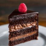 A close-up shot of a rich, layered Dark Chocolate Mousse Cake with raspberry filling, topped with a fresh raspberry and glossy chocolate glaze.