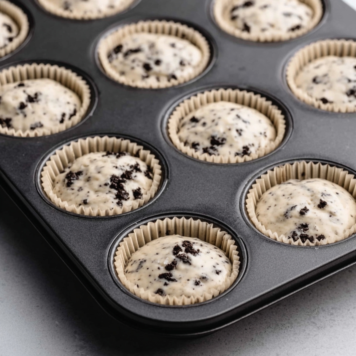 Delicious Cookies and Cream Cupcakes 11 Muffin tray filled with cookies and cream cupcake batter, topped with crushed Oreo pieces before baking.