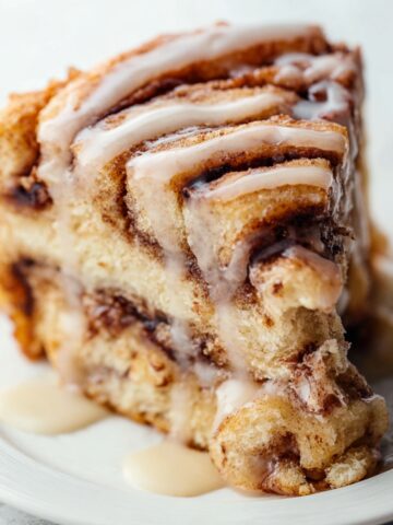 Easy Giant Cinnamon Roll Cake Recipe 7 Slice of Giant Cinnamon Roll Cake with cinnamon swirls and vanilla icing dripping down the sides on a white plate.