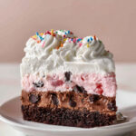 Slice of layered Ice Cream Cake Recipe with brownie base, chocolate layer, strawberry ice cream, and whipped cream topping