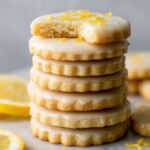 A stack of beautifully glazed Lemon Shortbread Cookies, with a bite taken out of the top one, showing the soft, buttery interior.