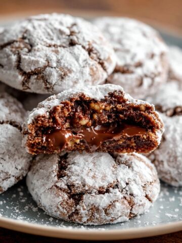 Soft Nutella Crinkle Cookies Recipe 7 A close-up of freshly baked Nutella Crinkle Cookies with powdery sugar coating, showing a soft and gooey interior with melted Nutella and hazelnuts.