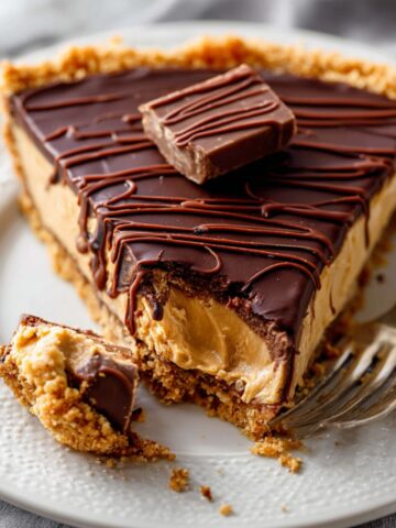 Peanut Butter Cup Tart 22 A slice of Peanut Butter Cup Tart on a white plate, with a bite taken out, showing the smooth peanut butter filling and chocolate topping. A fork rests next to the slice.