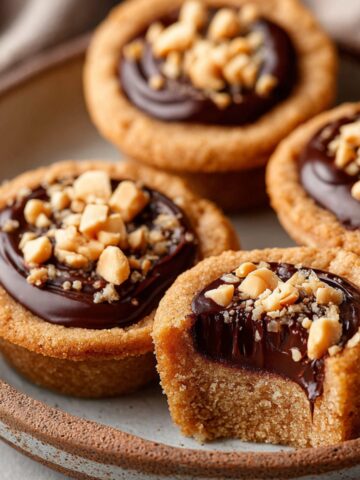 Peanut Butter Fudge Puddles Recipe | Easy Cookie Cups 8 Peanut Butter Fudge Puddles filled with chocolate fudge and topped with chopped peanuts on a plate, one cut open showing the gooey center