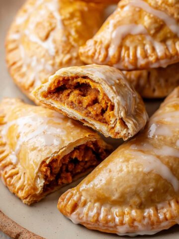 Pumpkin Hand Pies Recipe 16 Pumpkin Hand Pies arranged on a plate, with a bite taken out of one, revealing the spiced pumpkin filling inside. The pies are glazed with a light shine, showcasing their crisp, flaky texture.