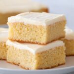 Soft vanilla sheet cake squares with smooth buttercream frosting stacked on a plate, showing a moist and fluffy crumb texture.