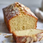 A freshly baked Almond Pound Cake with a light golden-brown crust, topped with slivered almonds and a dusting of powdered sugar. The cake is sliced to reveal its fluffy, moist texture inside.