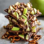 A close-up image of homemade Caramel Apple Bark With Pretzels topped with pretzels, chopped nuts, and green apple pieces. The bark is drizzled with extra chocolate, making for a visually appealing and sweet treat stacked neatly on parchment paper.