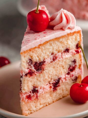 Easy Cherry Chip Cake Recipe 2 A slice of Cherry Chip Cake with pink frosting, topped with maraschino cherries, placed on a plate.