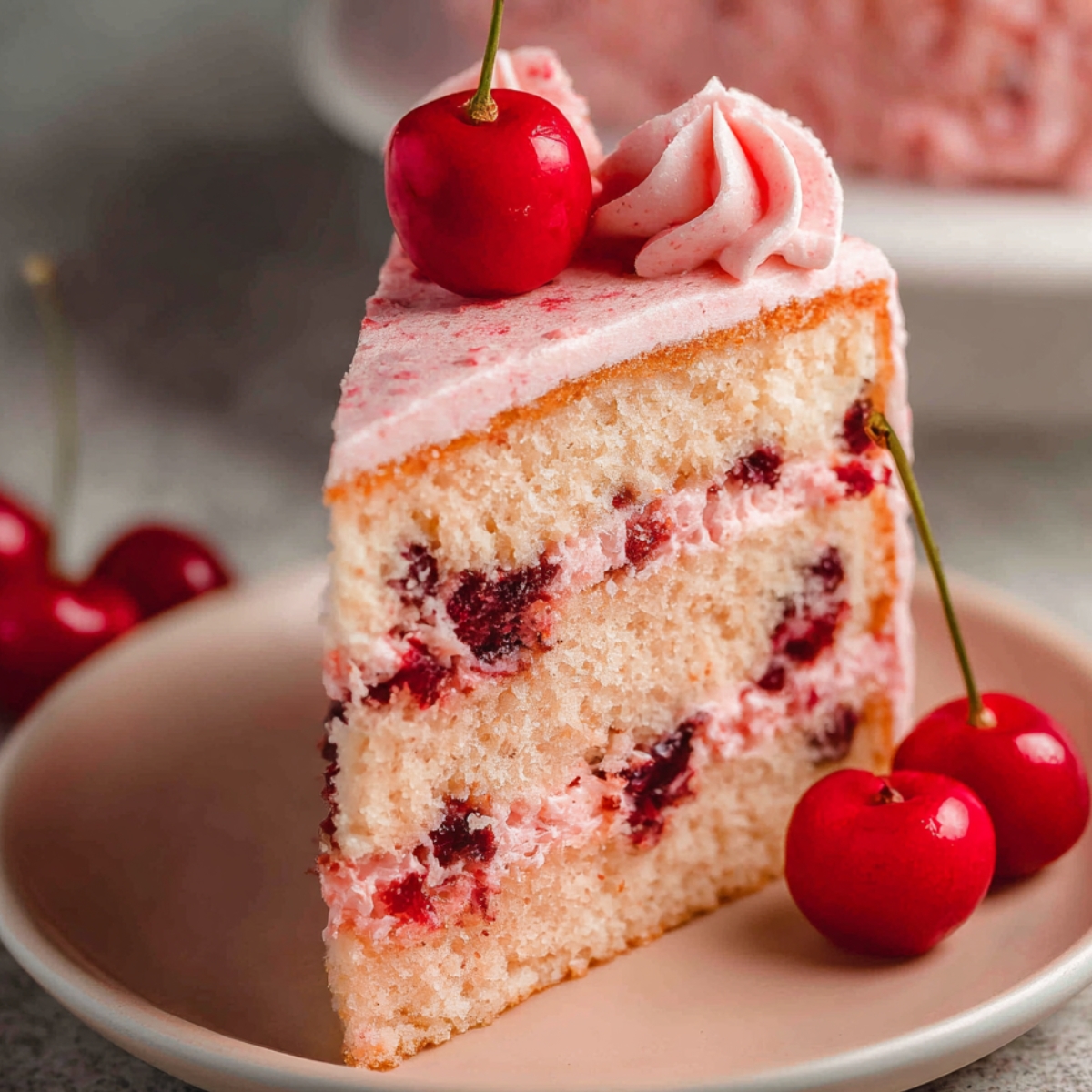 A slice of Cherry Chip Cake with pink frosting, topped with maraschino cherries, placed on a plate.