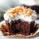 A freshly baked Chocolate Coconut Cupcakes with a bite taken out, revealing the rich, moist texture inside. The cupcake is topped with a layer of fluffy white frosting and toasted coconut flakes.