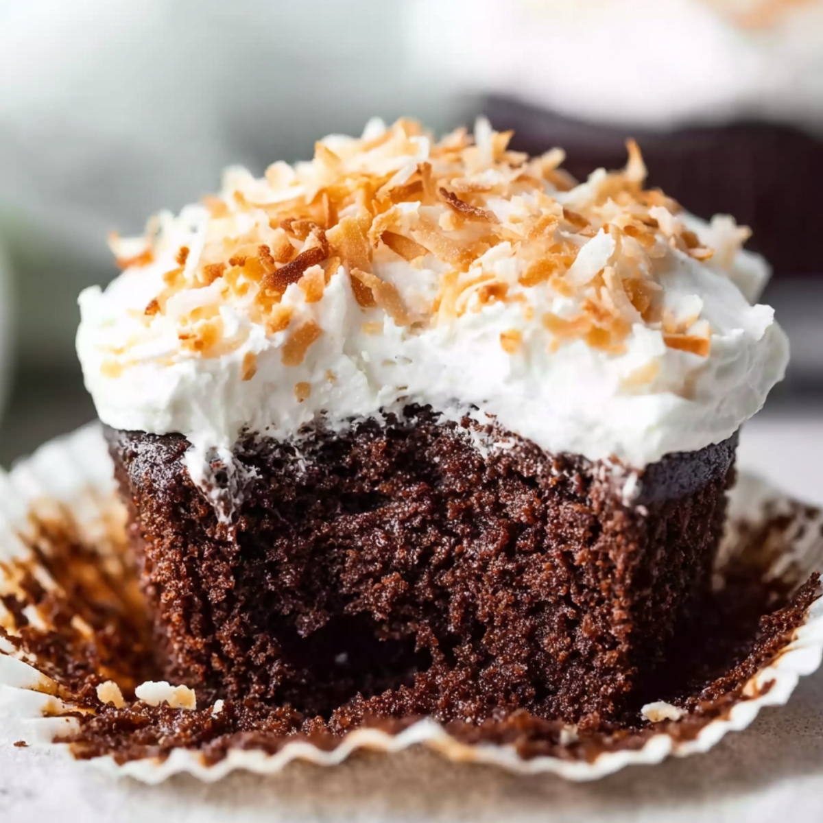 Moist Chocolate Coconut Cupcakes Recipe 9 A freshly baked Chocolate Coconut Cupcakes with a bite taken out, revealing the rich, moist texture inside. The cupcake is topped with a layer of fluffy white frosting and toasted coconut flakes.