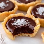 A close-up of a Chocolate Tartlets filled with smooth ganache, topped with a sprinkle of sea salt, with a bite taken out showing the creamy interior.