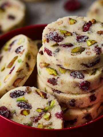 Cranberry Pistachio Shortbread Cookies 1 Stacked Cranberry Pistachio Shortbread Cookies with a sugar coating, arranged in a red container.