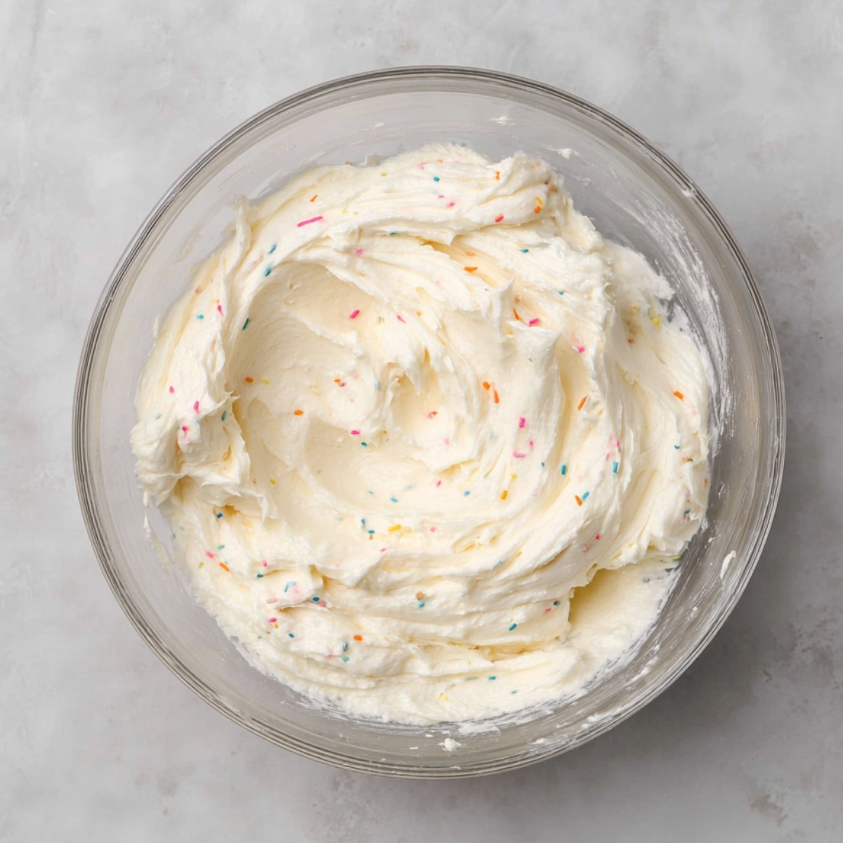 Funfetti Cookie Sandwiches Recipe 11 A glass mixing bowl filled with fluffy buttercream frosting, speckled with colorful sprinkles. The frosting is smooth and creamy, ready to be used for decorating.