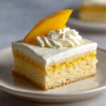 A slice of Mango Tres Leches Cake, topped with whipped cream and a fresh slice of mango, showcasing the layered texture.