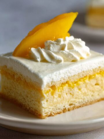A slice of Mango Tres Leches Cake, topped with whipped cream and a fresh slice of mango, showcasing the layered texture.