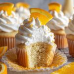 A cut Mimosa Cupcakes showing a soft and fluffy inside, topped with whipped cream, orange zest, and an orange slice.