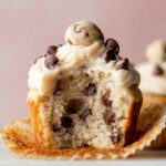 A freshly baked Molten Cookie Dough Cupcakes, topped with smooth buttercream frosting, mini chocolate chips, and a ball of cookie dough. The cupcake is partially unwrapped to reveal the soft interior filled with chocolate chips.
